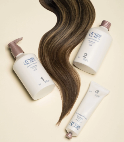 Ochre brand hair care products including a purifying shampoo, dermal conditioner, and hair cream, arranged around a long, wavy strand of brown hair on a beige background.