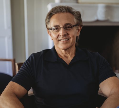 Shawn Khorrami cropped Shawn Khorrami smiling while seated indoors, wearing a dark navy polo shirt and glasses, with a fireplace and light walls in the background.