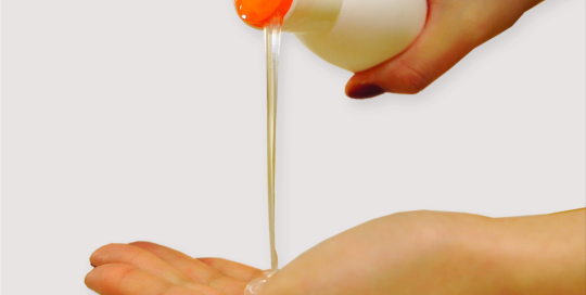 Clear liquid pours from an orange disc top cap held by two hands over an open palm.