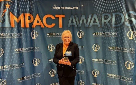 Coni Lefferts smiles and proudly holds her award in front of the Impact WBEC background.