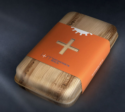Orange Cross Wood A rectangular wooden soap box with rounded edges is wrapped with an orange paper band featuring a cut-out cross symbol. The wood grain is clearly visible through the smooth finish, and the orange label contrasts strongly with the light wood. The design suggests a natural cosmetic product with modern branding.