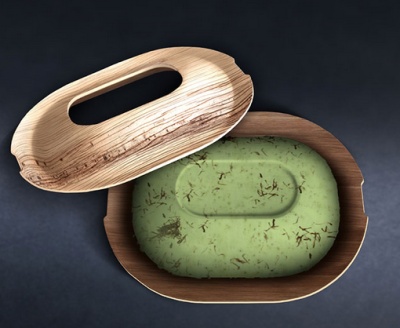 Palm Leaf Soap A green oval bar of soap rests inside a shallow, oval soap dish made from pressed palm leaf. The soap contains visible dried plant pieces embedded throughout. A matching palm leaf lid is placed slightly open beside the dish, showing the soap inside. The overall look is simple, natural, and eco-friendly.