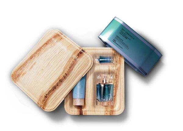 An open square cosmetic box made from pressed palm leaf shows three blue skincare items arranged inside molded compartments: a small tube, a pump bottle, and a small vial. The matching palm leaf lid is placed slightly to the left of the box, showing the natural wood grain and layered texture. A larger blue cylindrical container rests outside the box at the upper right. The overall presentation emphasizes natural materials paired with sleek blue cosmetic packaging.