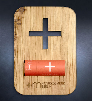 Palm Leaf Wood With Cross A flat wooden display card features a large cross-shaped cutout near the top. Below the cutout, a small orange lip balm tube with a white cross logo is attached horizontally. The wood surface shows natural grain and engraved branding text, reinforcing a minimalist, natural cosmetic presentation.
