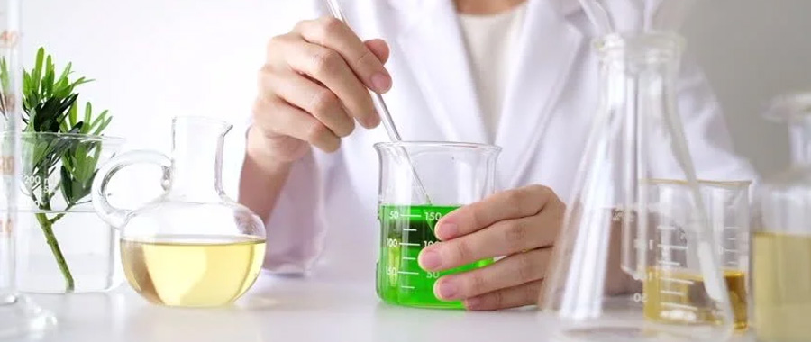 header-for-jennifer-di-marco A scientist in a lab coat mixing a green beaker on a table. There are two yellow beakers on either side of the green beaker.